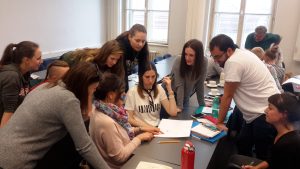 Various persons sitting and standing around a table, all looking at a piece of paper. 1 person is pointing on it.