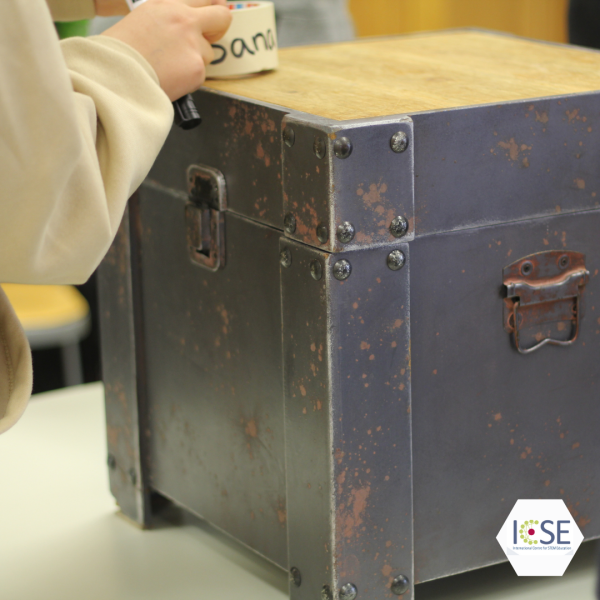 A metal-wooden escape box with one hand on top of it.