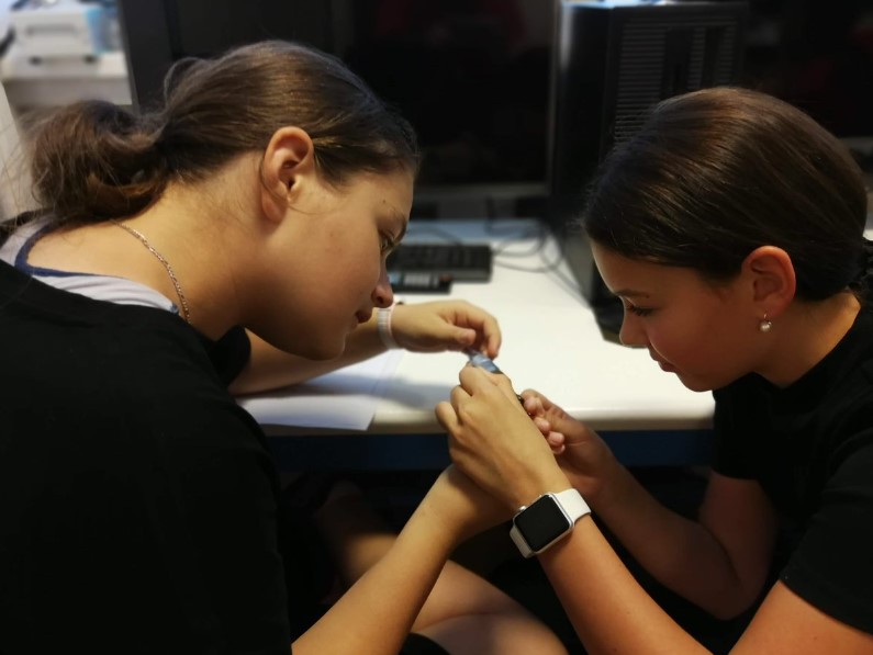 Two schoolgirls are working together on something in their hands. They are sitting facing each other and are in a computer room.
