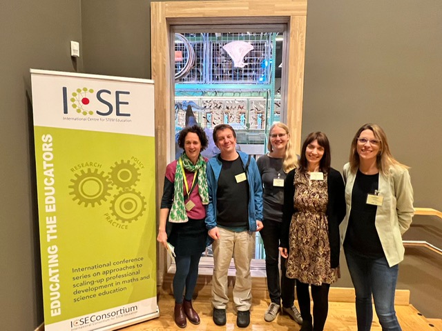 5 Team members of the ICSE Team stand next to the green Educating the Educator Banner and smile into the camera.