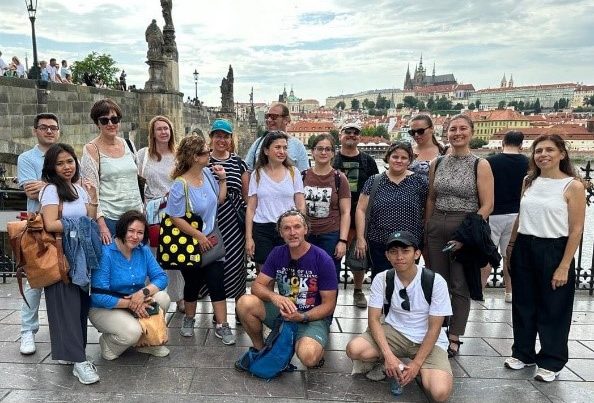 A group of summer school students are standing in front of the city skyline of Prague. There are a total of 17 individuals, some kneeing on the floor, others standing in the back. There are some clouds with blue sky, shining through them.