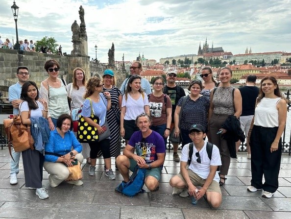 A group of summer school students are standing in front of the city skyline of Prague. There are a total of 17 individuals, some kneeing on the floor, others standing in the back. There are some clouds with blue sky, shining through them.