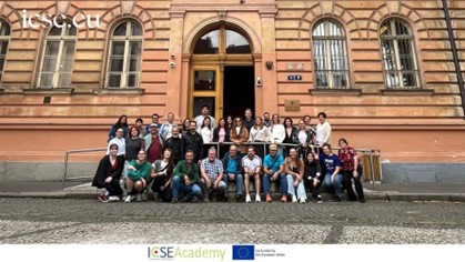 A group of about 30 people is sitting and standing on the stairs of a red historic building.