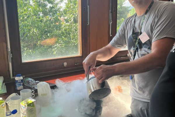 A person is puring something from one pot into another one. There is a lot of fog all around the pots. He is wearing a grey shirt and grey pants. On the table are various utensiles, like spoons, a cardboard jug, a water bottle and tea cups. In the background there is a window with green plants and a blue sky just outside of it.