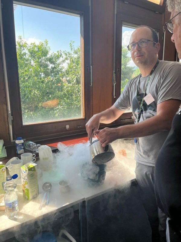 A person is puring something from one pot into another one. There is a lot of fog all around the pots. He is wearing a grey shirt and grey pants. On the table are various utensiles, like spoons, a cardboard jug, a water bottle and tea cups. In the background there is a window with green plants and a blue sky just outside of it.