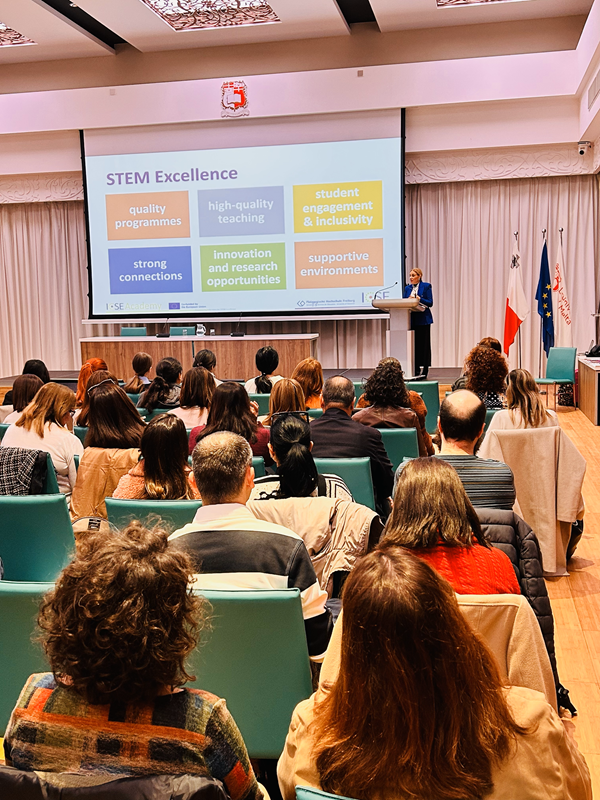 A person gives a presentation inan auditorium to an audience. The headline of the presentation is "STEM Excellence"