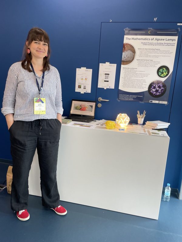 A brown and long haired person is standing in front of a white booth in front of a blue wall. Behind the booth is a poster of the ICSE Factory project. On the booth there are a variety of items, amongs them a brightly lit up and interestingly shaped white lamp and pens.