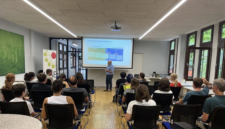 A person standing in front of a room filled with people sitting in black chairs. The person is standing in front of a white wall with a projection on it. On the left side there is a Banner with the ICSE C.