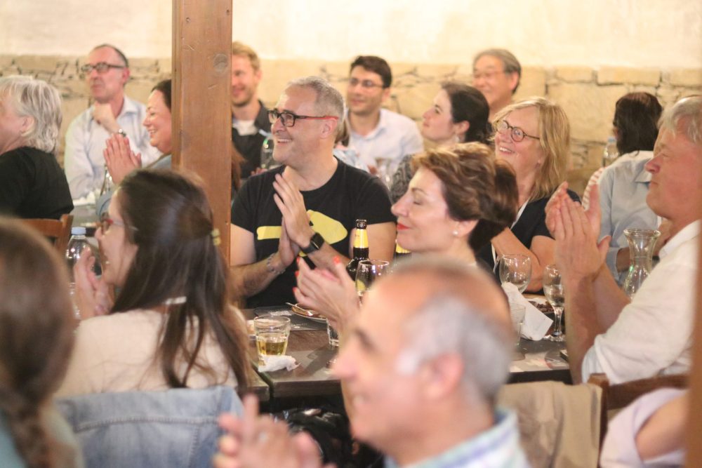 A group of people are sitting on wooden tables in a restaurant and are clapping an laughing and looking towards one side of the room.