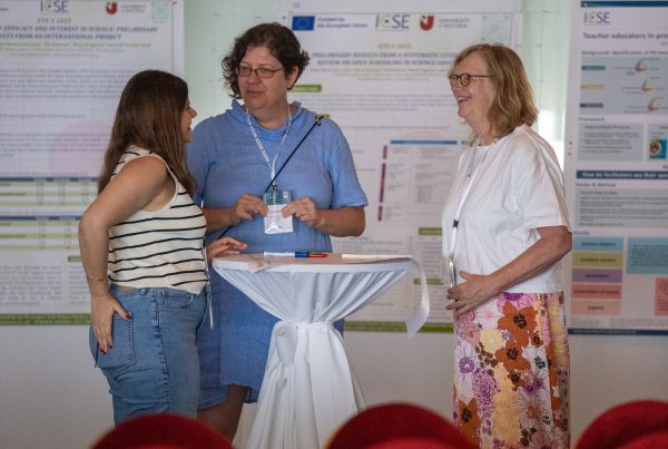 3 people are standing around a table talking and smiling. In the background here are posters from various projects
