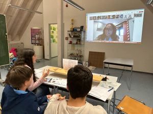 A teacher and two students sit at a classroom table with worksheets and a small experiment setup, participating in a live video call projected on the wall with a partner class in another country as part of an international school collaboration.
